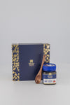 Manuka Honey gift set featuring a navy blue box with gold floral borders, a small jar of colonics honey with a blue lid, and a wooden spoon on a light gray background.