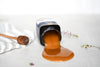 Honey drips from an open jar onto a white surface with a wooden honey dipper nearby.